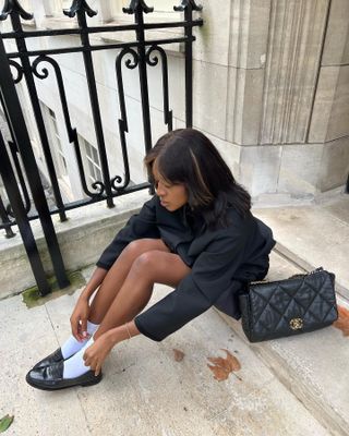 Influencer sitting down on marble steps, wearing black jacket, black Chanel back, and adjusting white socks while wearing black loafers.