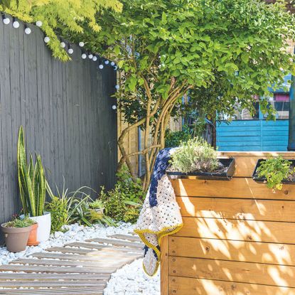 Garden area with a wooden decked path surrounded by stones and plants growing up the black painted fence