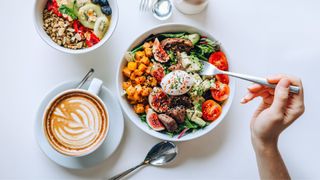 Bowl of salad with tomatoes, avocados, egg, figs, and mango next to bowl of granola and flat white coffee