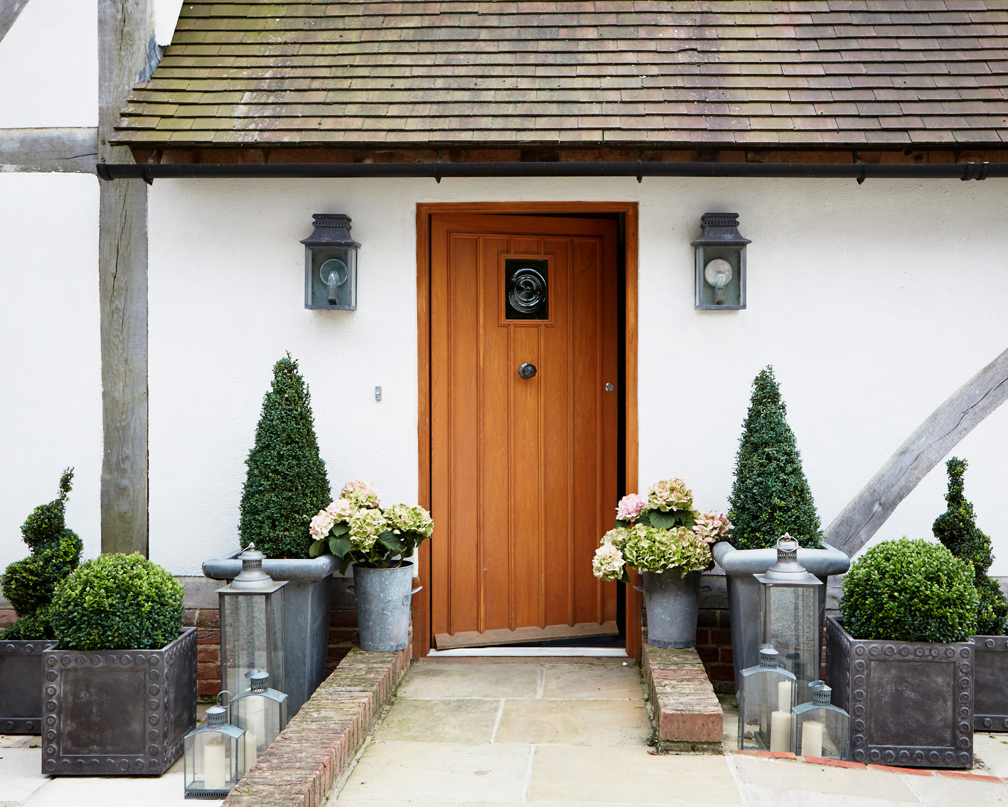 An example of how to decorate a front porch showing a white house with a wooden front door surrounded by plant pots