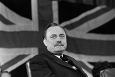 Conservative Party politician Enoch Powell seated in front of a British Union Jack flag
