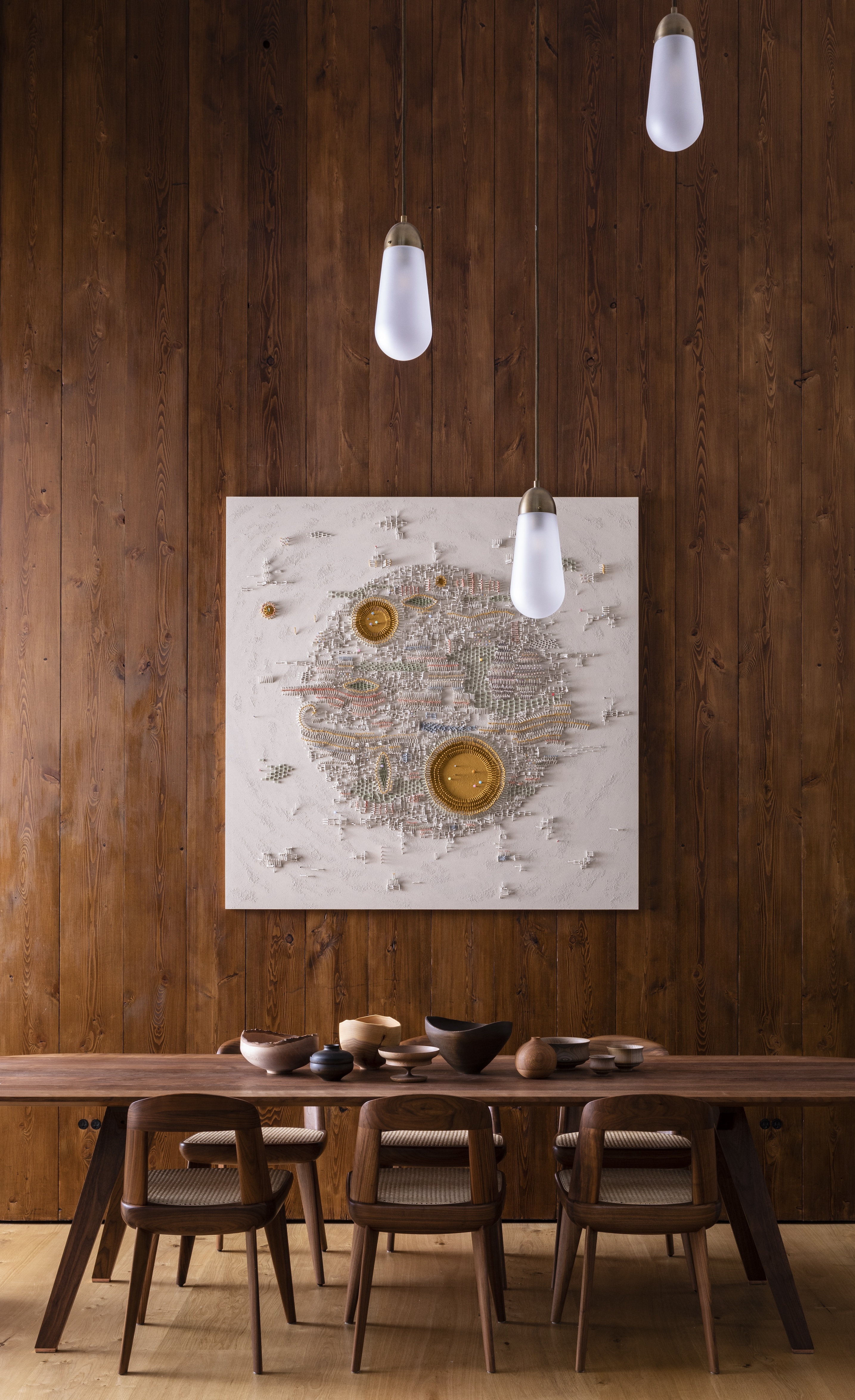 A wooden dining table in front of a wood paneled wall with a large textured artwork and tear-drop pendant lights hanging down