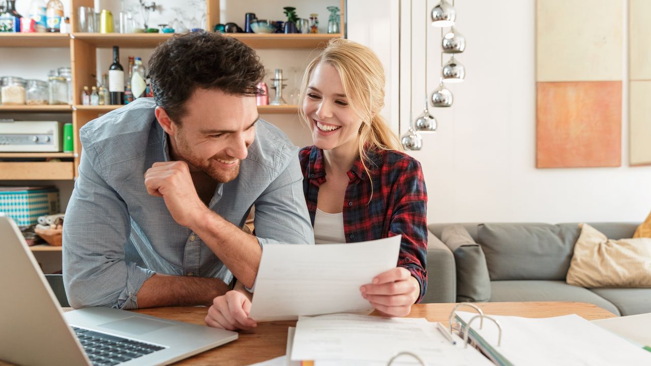 A young couple smile as they look over their spending paperwork at their dining room table.