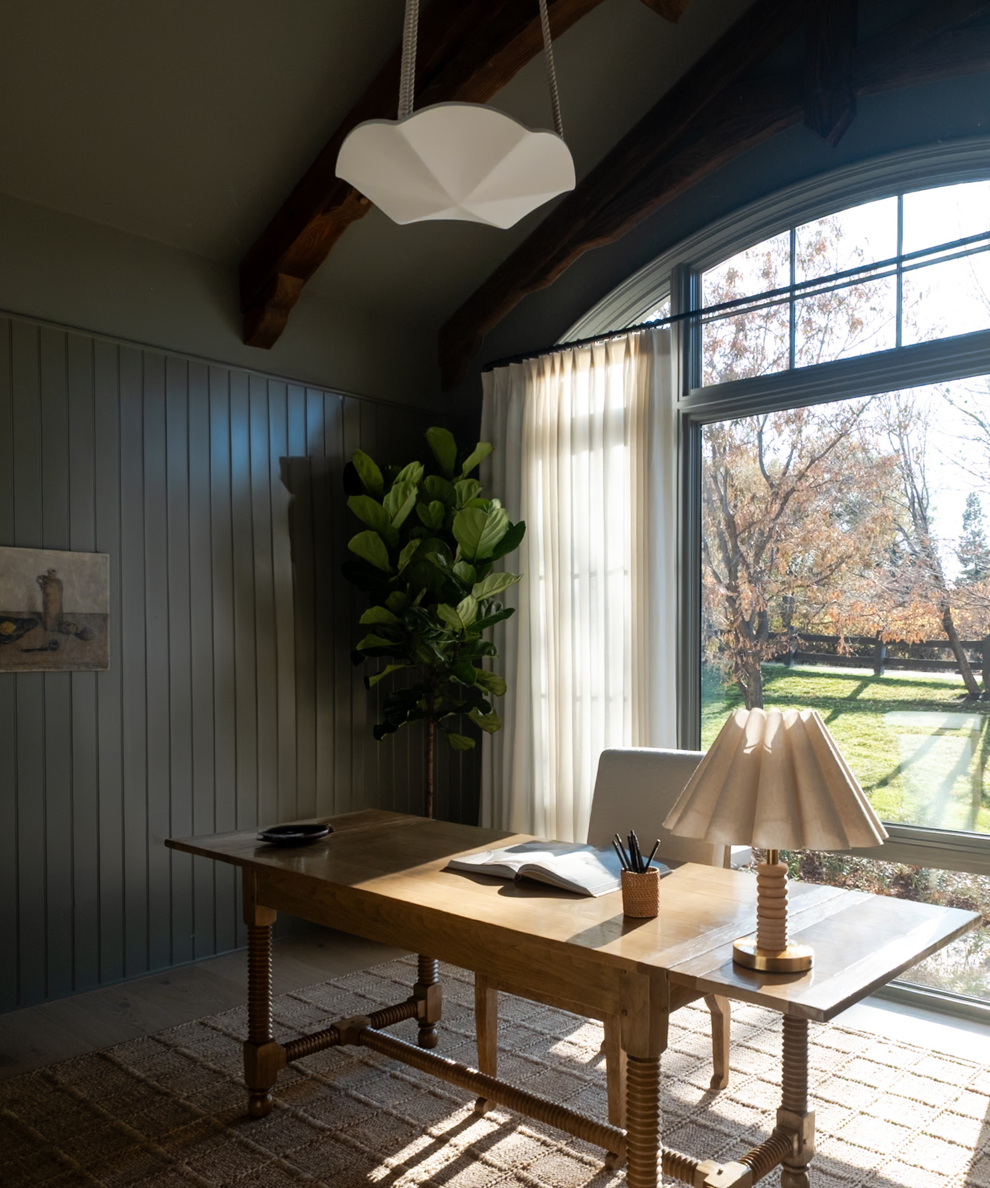 a room with vaulted ceilings, a large window, a large wood desk, and dark painted walls