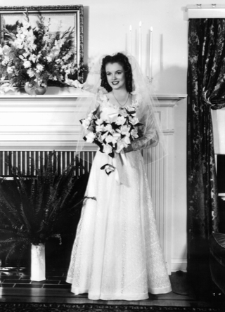 Marilyn Monroe, on the day of her wedding day to James Dougherty, in front of a fireplace in her wedding dress and holding a bouquet, Los Angeles, 1942