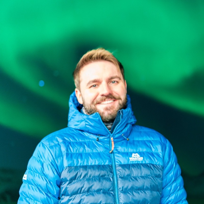 a man wearing a blue coat smiling with green aurora in the background.