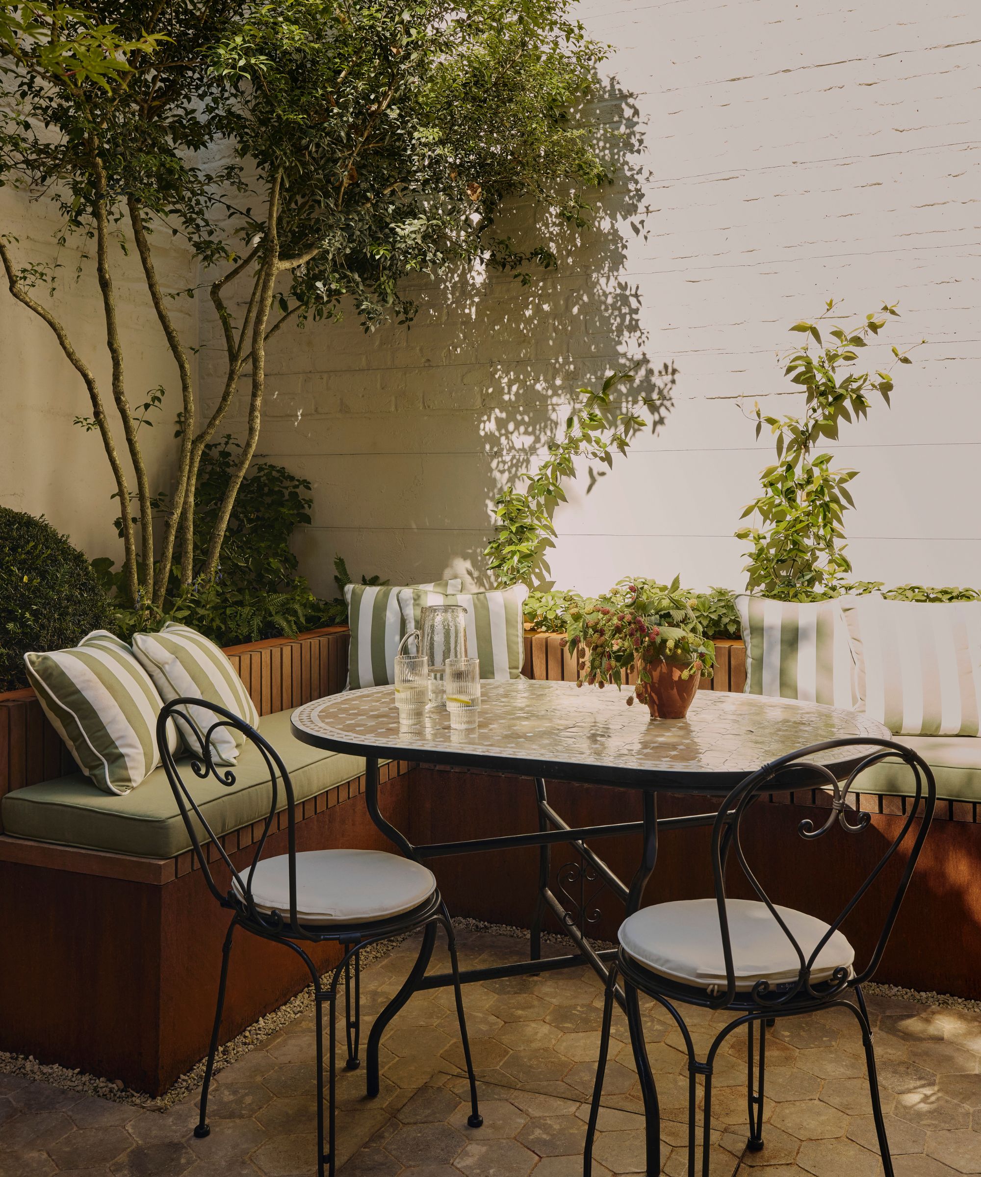Outdoor patio with banquette seating, wrought iron table and chairs, striped cushions and potted tree in the corner