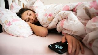 Teenage girl sleeping on side in bed with pink and white patterned bedding next to mobile phone