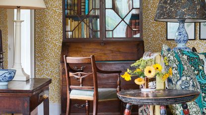 An antique-filled living room featuring yellow and white Morris & Co. "Willow Bough" patterned wallpaper. In the center is a dark wood secretary desk with a glass-front bookshelf filled with old books, paired with a matching wooden armchair. To the right, a round side table holds a small vase of bright yellow flowers, set against a backdrop of vibrant green and teal floral patterned cushions. Two large decorative lamps with patterned shades frame the scene, and various blue and white porcelain pieces add classic detail to the warm, eclectic interior.