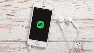 En mobil med ett par vita hörlurar som ligger mot ett ljust träfärgad bord, med Spotify-appen uppe på skärmen.