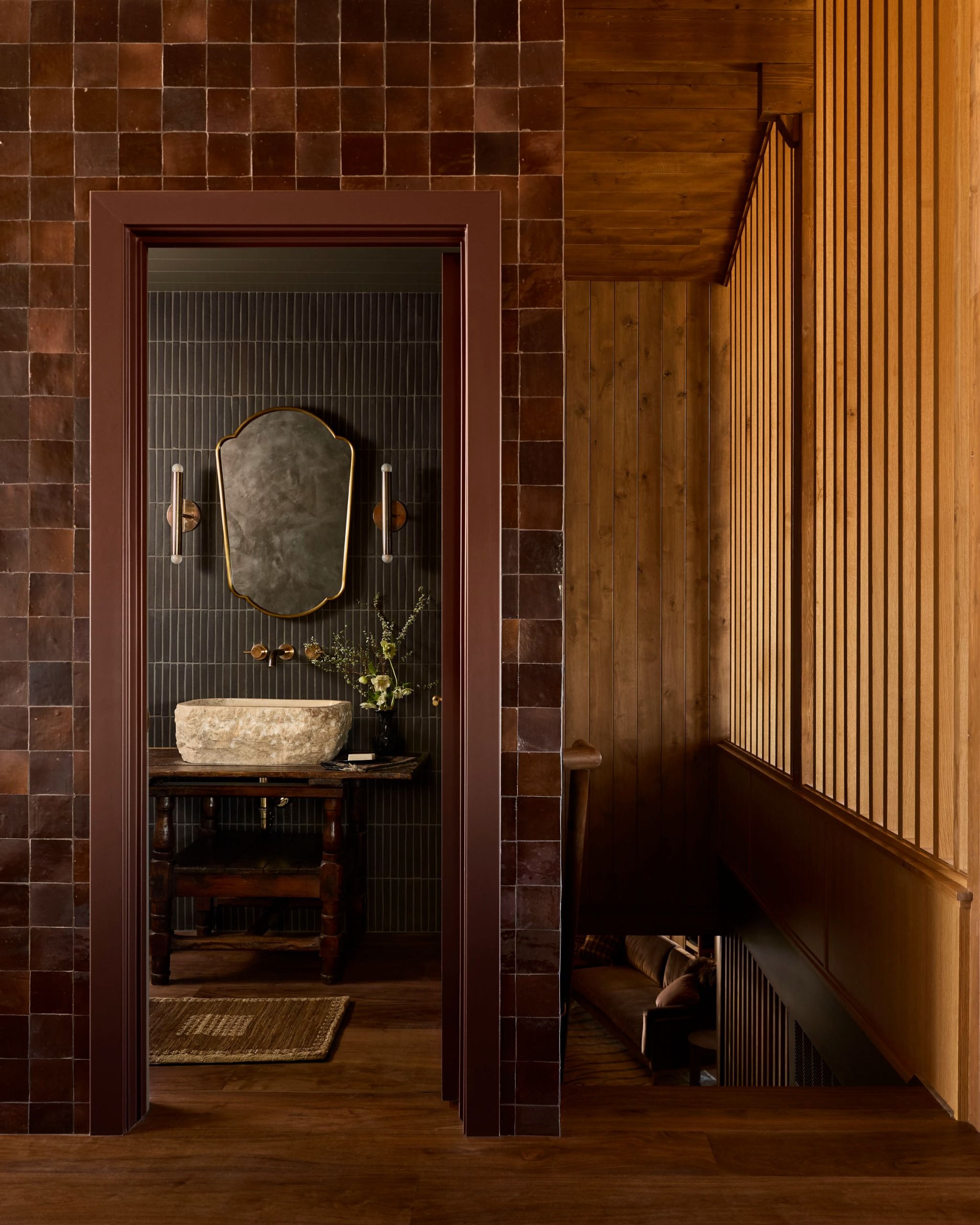 The doorway of a powder room clad in tiles with wood paneling to the side of the stairs on the right