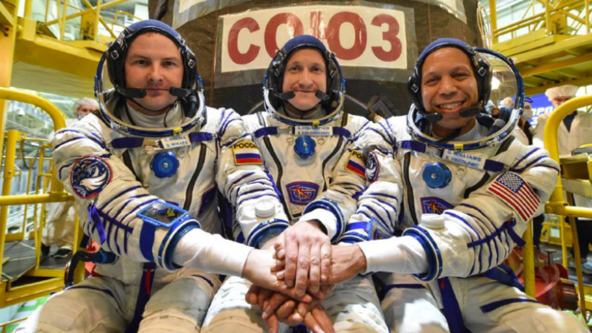 Three men clasp hands in Russian Sokol spacesuits ahead of the Soyuz MS-28 launch.