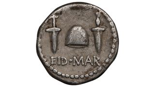 A silver coin against a white background. The coin has two daggers and a hat between them. It says EID MAR for the ides of march.