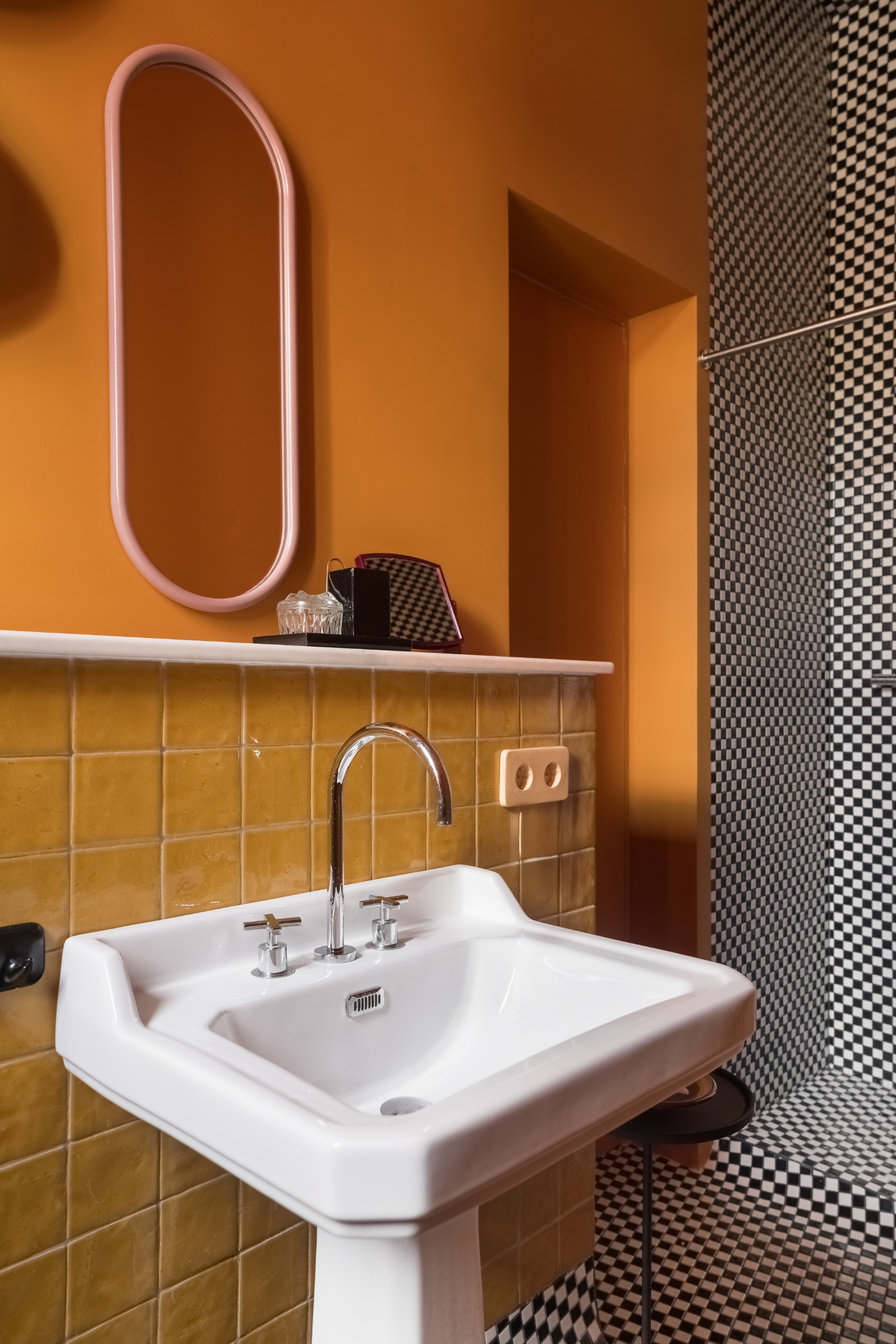 A bold bathroom with a an orange wall, yellow tiles, a while sink, a pink-framed oblong mirror and a small-tiled white and black checkerboard shower