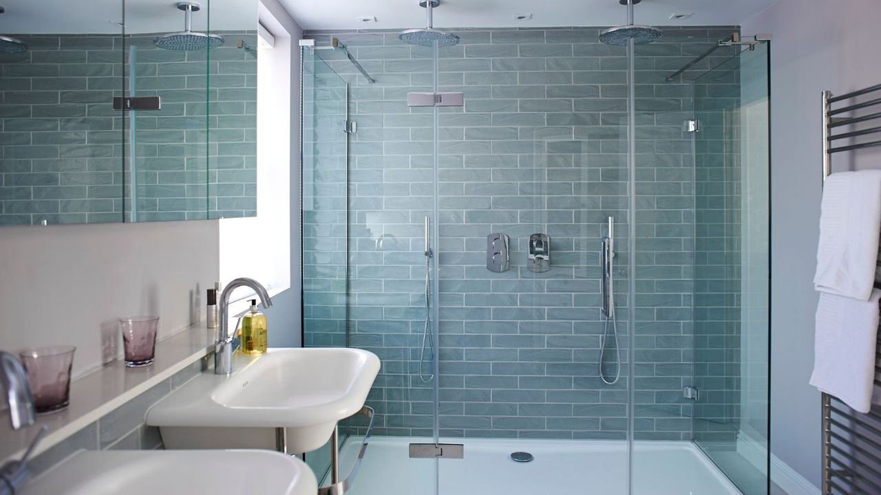 A blue tiled shower in a bright bathroom with a window, white matching sinks, purple glasses above with a yellow bottle of hand soap, and a silver radiator with two white towels hanging up