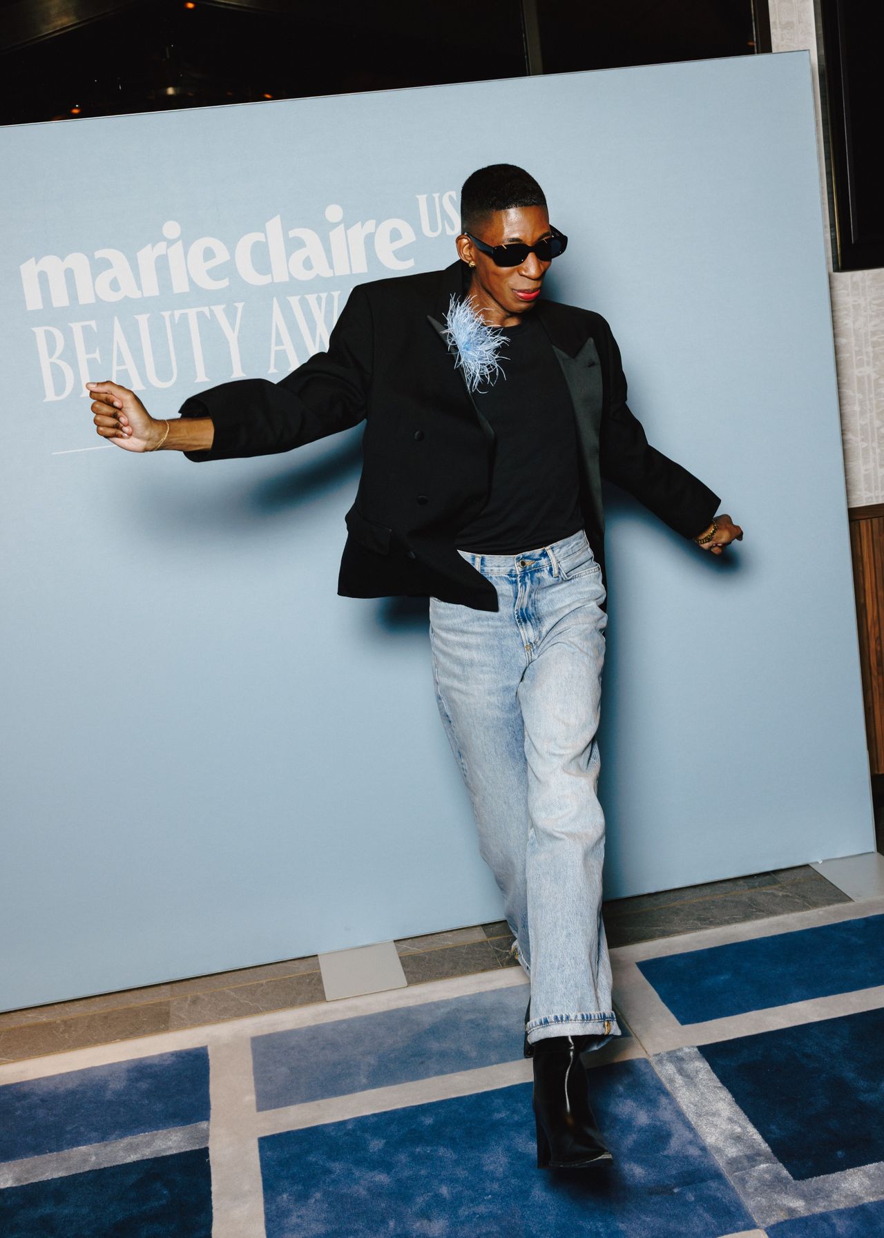 a black man in jeans and a blazer in front of a blue step and repeat