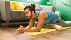 A man performs pigeon pose in a living room on a yoga mat in a vest and sweats. He is on the floor, with his right leg bent underneath his torso and his left leg extended behind. He leans forward on his elbows and smiles. Behind him we see a plant and a couch.