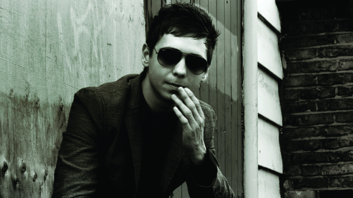 Laurence Jones sitting in front of a house, wearing shades, with his hand to his mouth