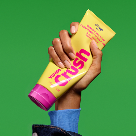 A hand holding a yellow and pink tube of lotion on a green background.