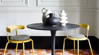 Wooden black round table with handled and ribbed pottery, yellow dining chairs with zebra print and black baseboards behind with white French-look paneled walls