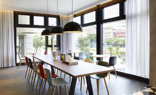 Dining area with large windows, long wooden table with multiple colored chairs and black hanging ceiling lights