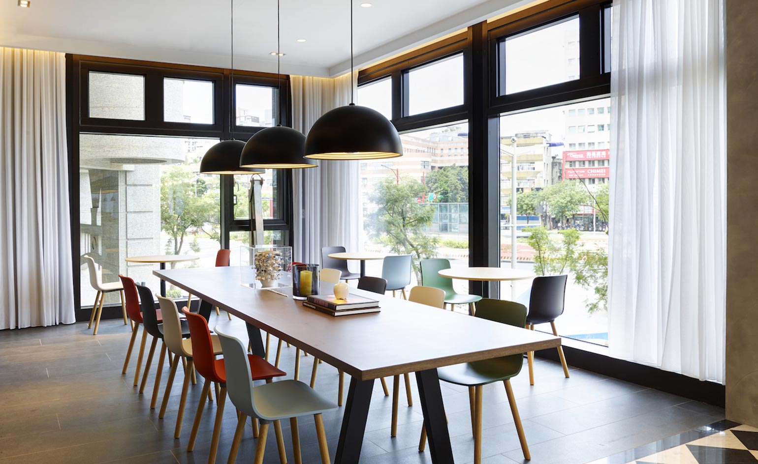 Dining area with large windows, long wooden table with multiple colored chairs and black hanging ceiling lights