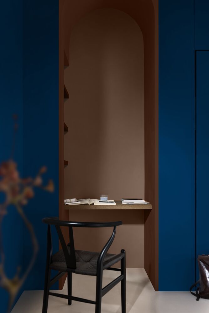 Image of a desk painted in a mushroom brown color with dark blue walls on either side. There is a black desk chair pulled up to the desk.