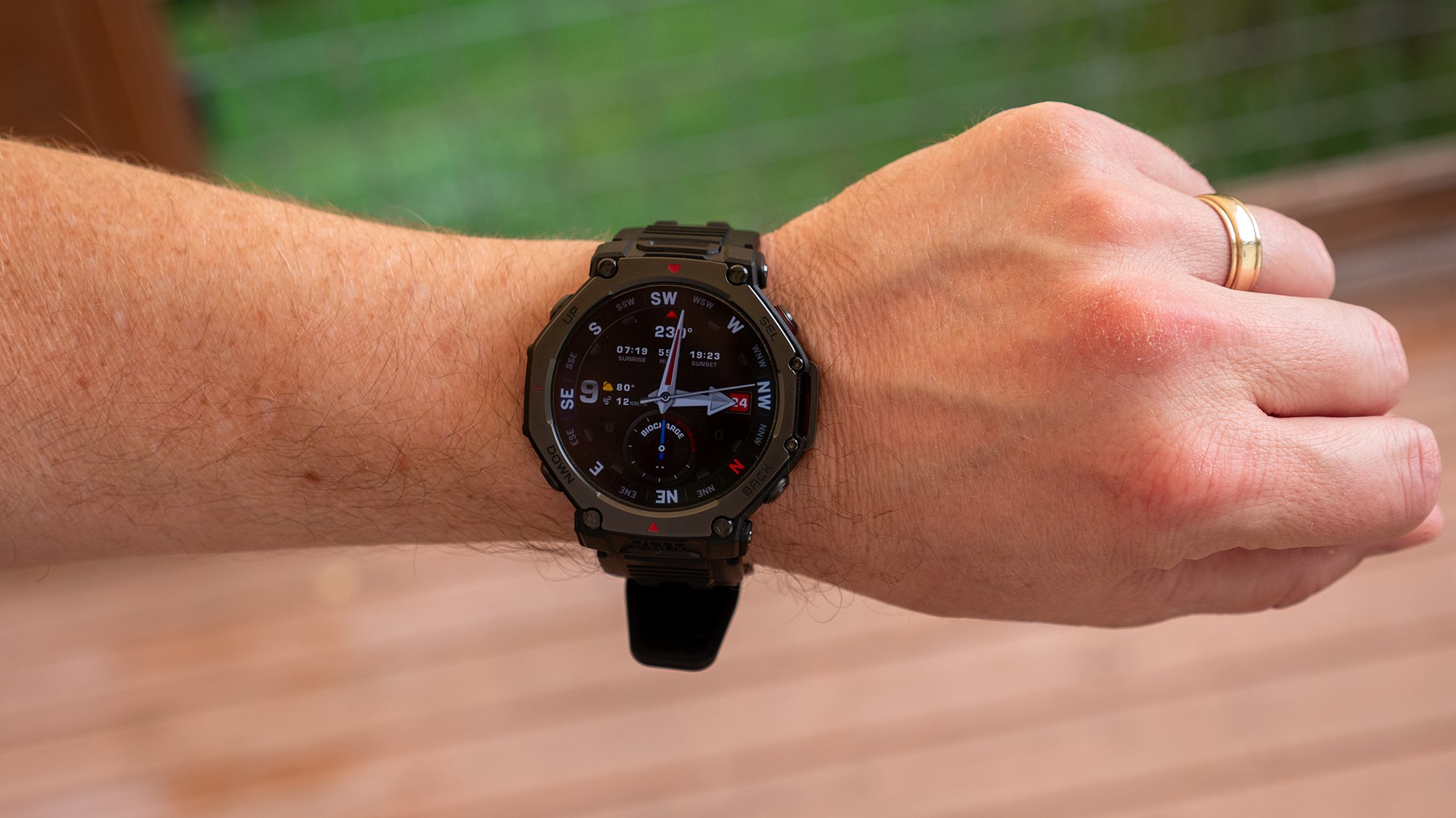 A photo of the Amazfit T-Rex 3 Pro on a man's wrist above a wooden deck, showing a standard watch face.