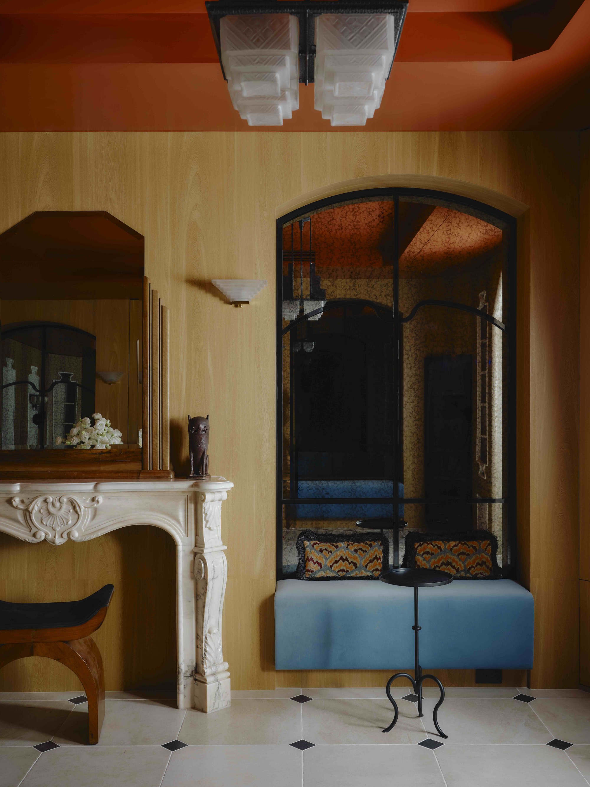 Image of a luxurious, Art Deco entryway with wooden walls, white tile floors, orange ceiling, and a bench seat built-in that has mirrored walls and a light blue velvet seat.