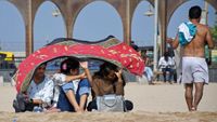 Three women sit on a beach in Mumbai, India, holding a cloth over their heads to protect themselves from the scorching sun.