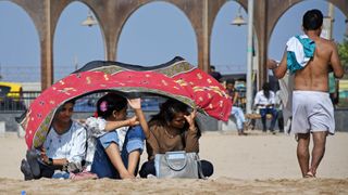 Three women sit on a beach in Mumbai, India, and hold a cloth over their heads to protect themselves from the scorching sun.