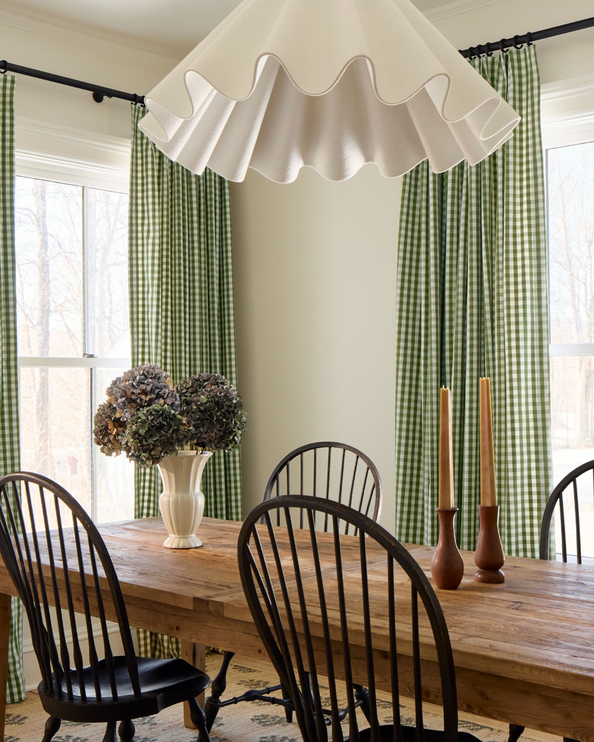 a bright, modern rustic dining room with two windows with green gingham cotton curtains, a rustic wooden dining table with four black spindleback dining chairs, and a wavy white pendant light