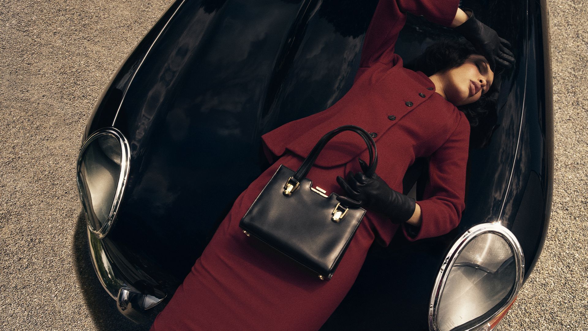 Nara Smith lays on a black vehicle while wearing a deep red suit and holding a handbag with her other hand over her forehead.