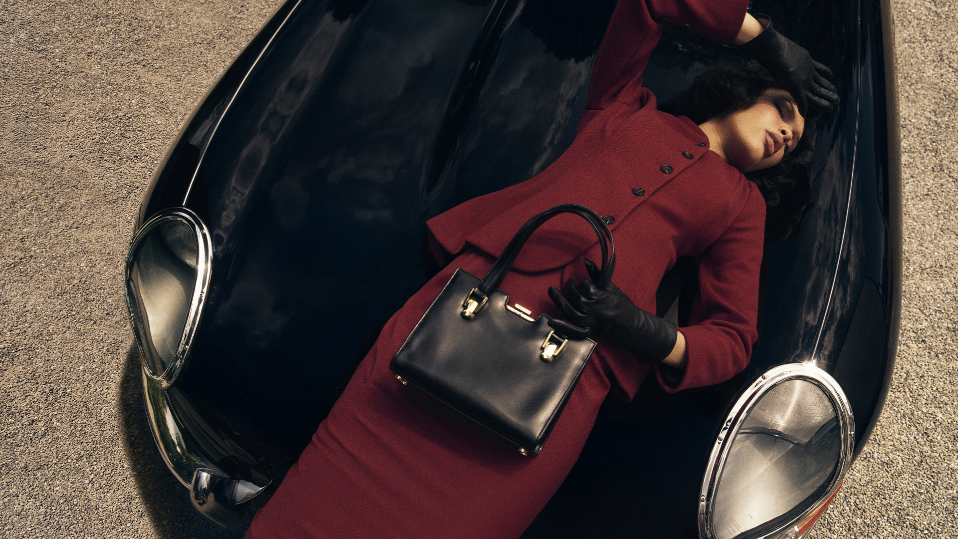 Nara Smith lays on a black vehicle while wearing a deep red suit and holding a handbag with her other hand over her forehead.