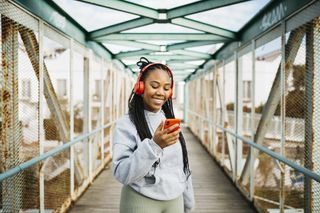 Portrait of a young smiling woman, with braided hair and sportswear, using a red smartphone and headphones, after workout on a urban bridge