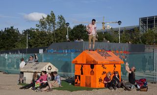 View of two orange and off-white coloured mini houses with various black designs all over. There is a man standing on top of the orange house and several people sitting on the ground close by. In the background there is fencing, hoarding, greenery and a building