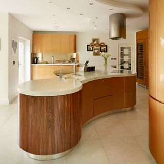 curved wooden island with white worktop, with wooden cabinets and stone coloured floor tiles