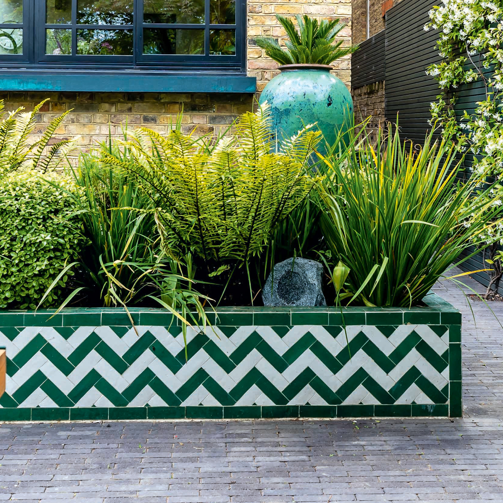 A raised bed planter covered in green and white chevron tiling packed with ferns and lush greenery