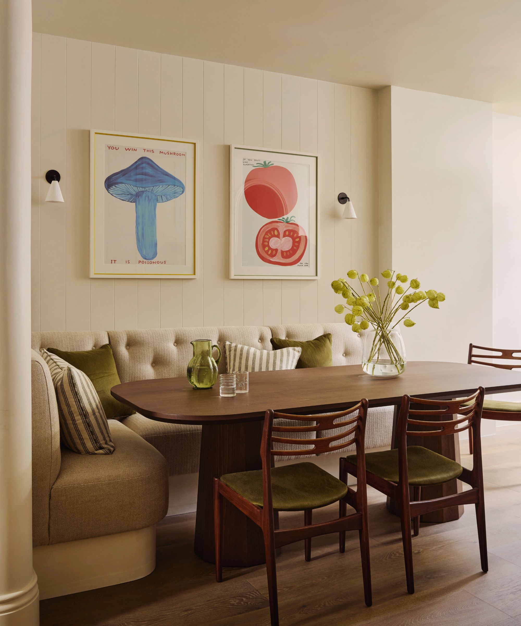 An open-plan dining area with a dark wood table, cream banquette seating, and produce-themed colorful artwork on the walls