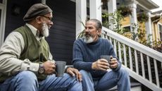 Two men sitting on a front porch talking and holding mugs