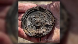 We see a hand holding the slightly corroded silver circular medal. There is dirt on it.