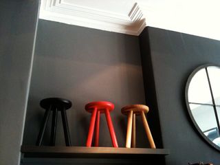 Three 3-legged wooden stools (left to right - black, red and brown) displayed on a black floating shelf photographed against black wall.