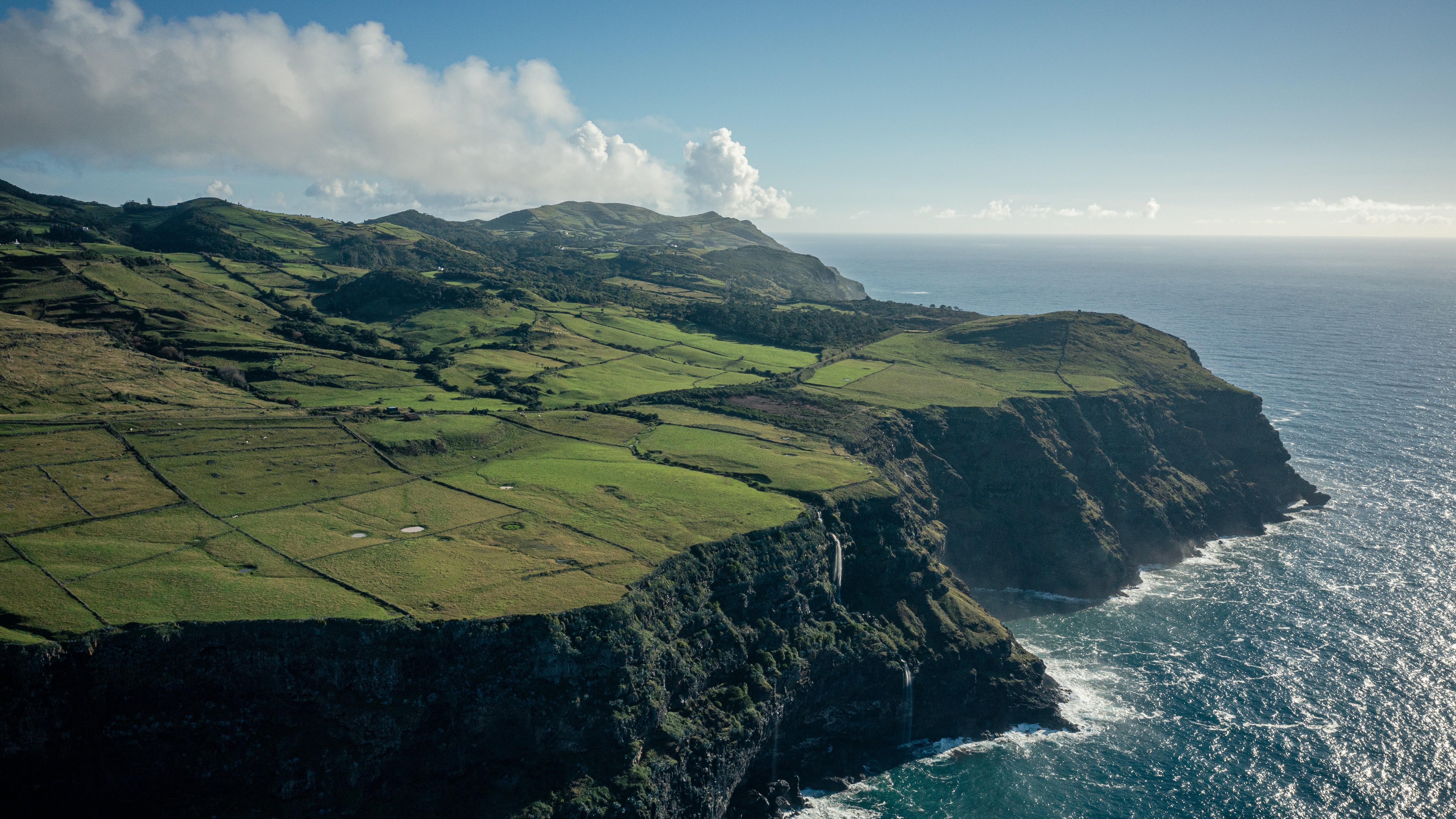 a blue ocean crashes against the bottom of a high cliff that gives way to green fields stretching to mountains in the distance.