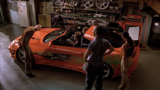 The main characters from The Fast and the Furious admiring the orange Toyota Supra in garage