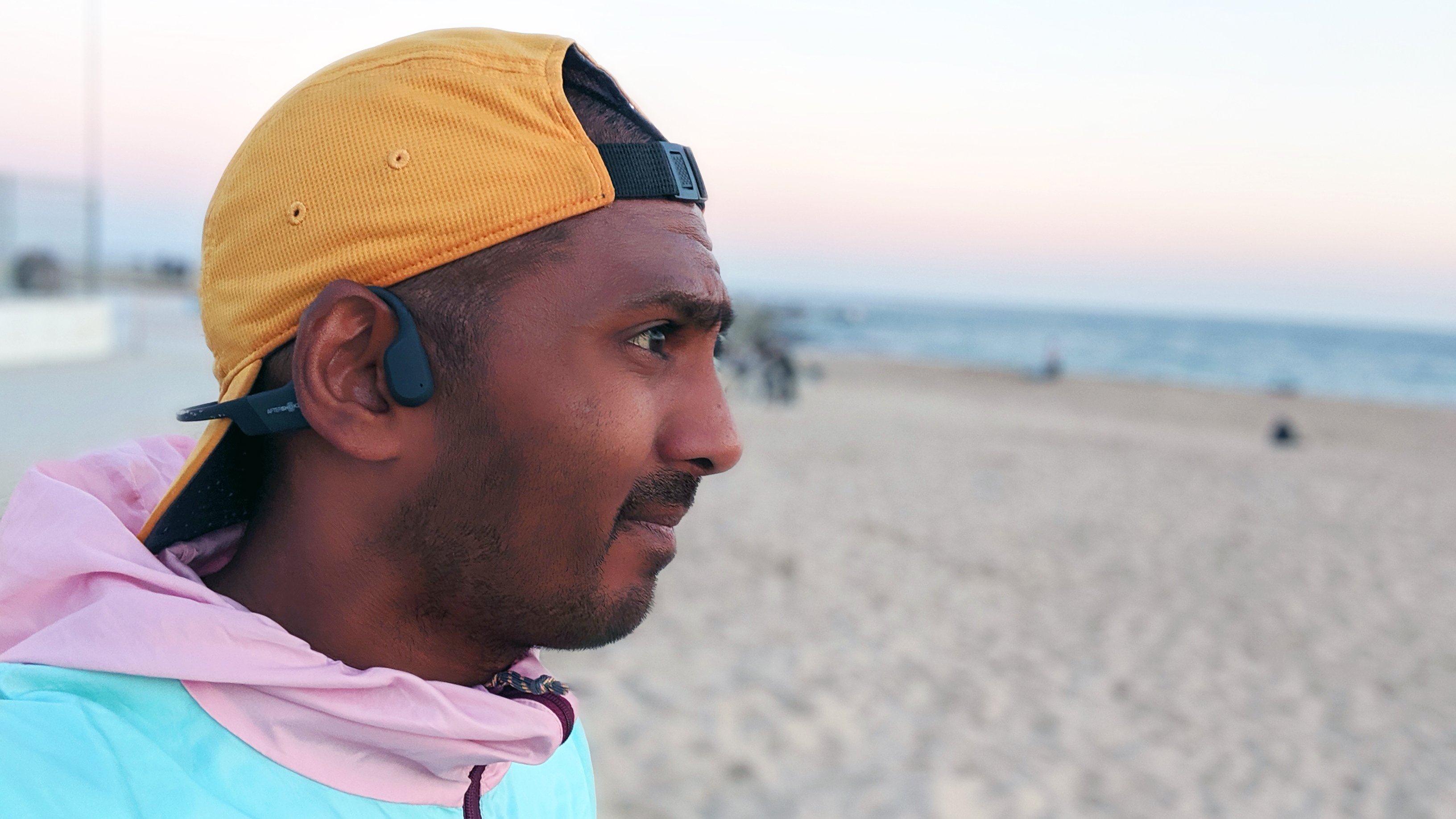 Penulis TechRadar Michael Sawh mengenakan headphone konduksi tulang Shokz OpenRun dengan pantai di latar belakang.