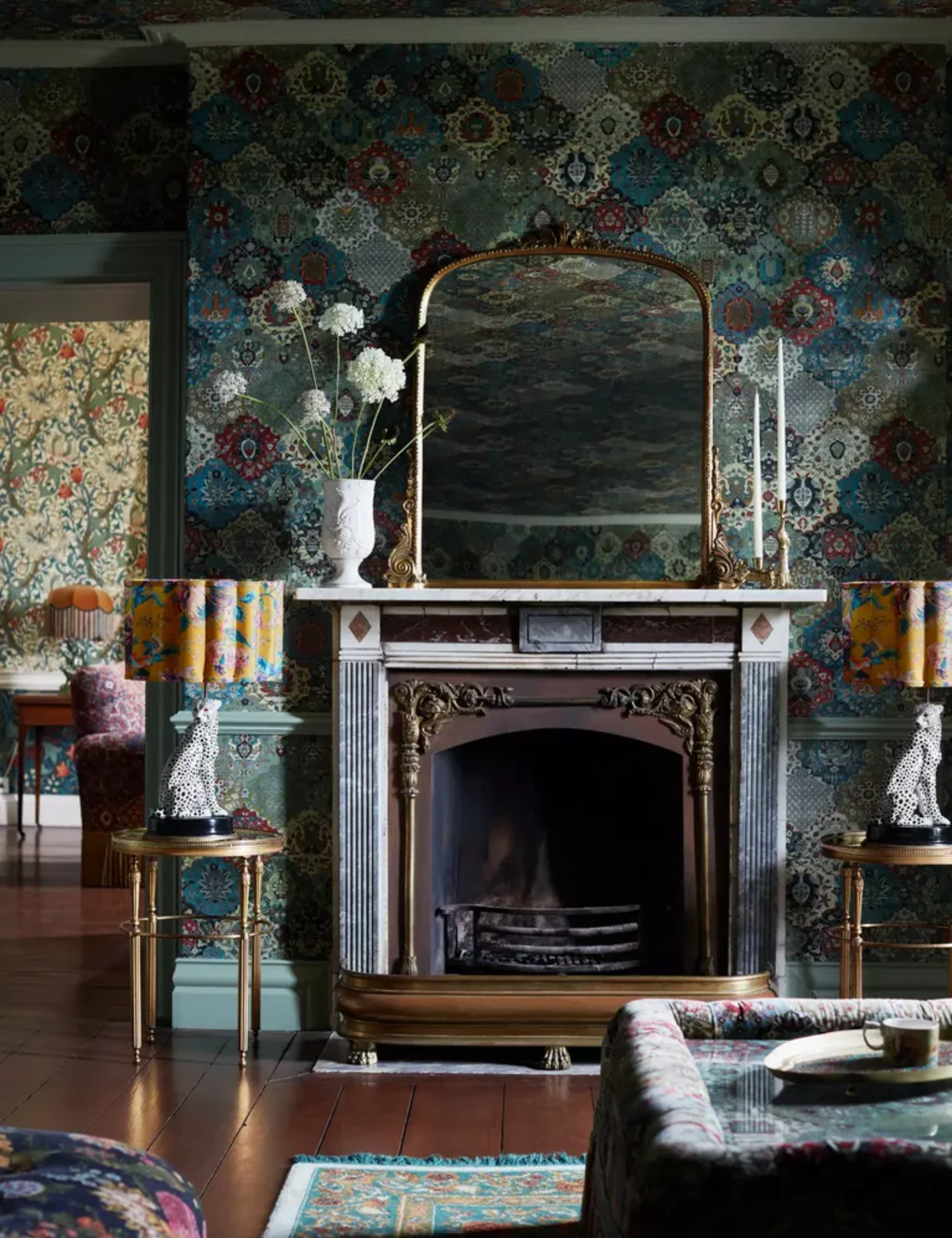 Anthropologie Gleaming Primrose mirror resting on the mantle of a wood and marble fireplace. Vignette is shot inside of a jewel-toned, living room covered in floral wallpaper.
