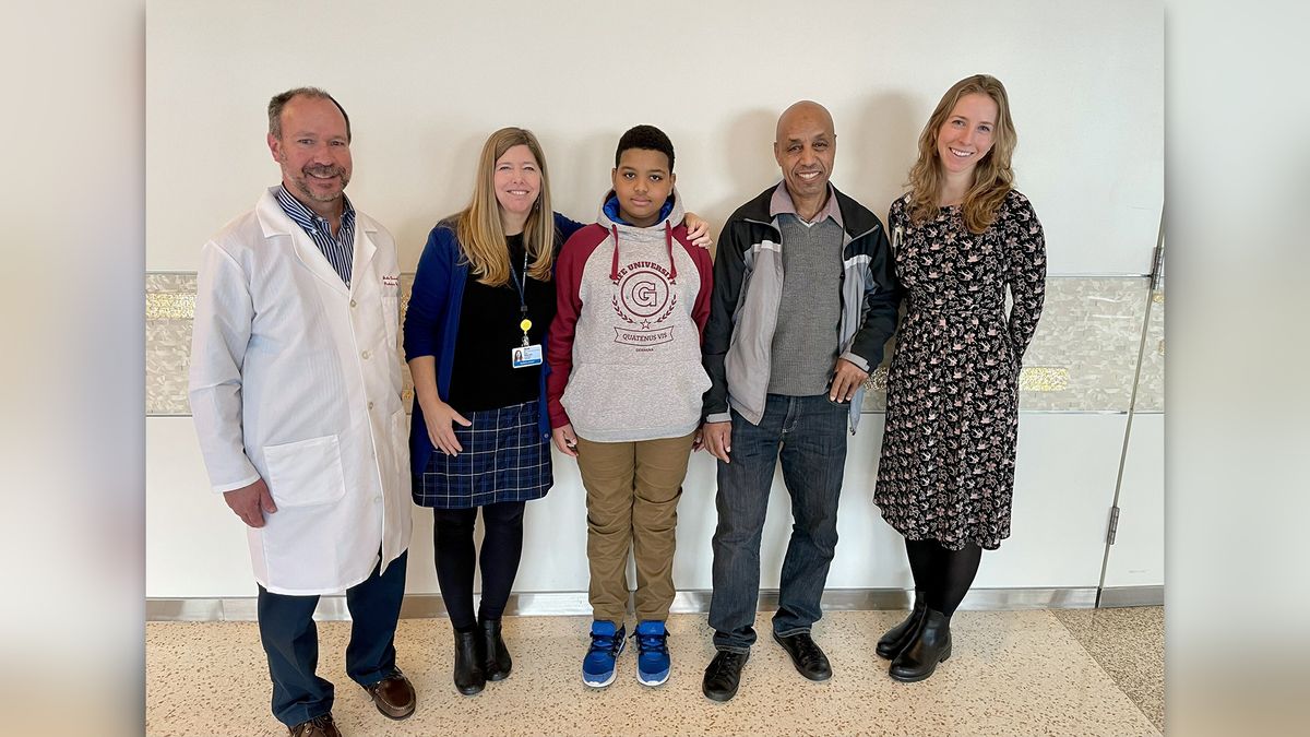 A photo of five people standing in a line: a male doctor in a white lab coat, a woman in professional clothing and a lanyard, a 11-year-old boy, the boy&#039;s father, and another woman in professional attire