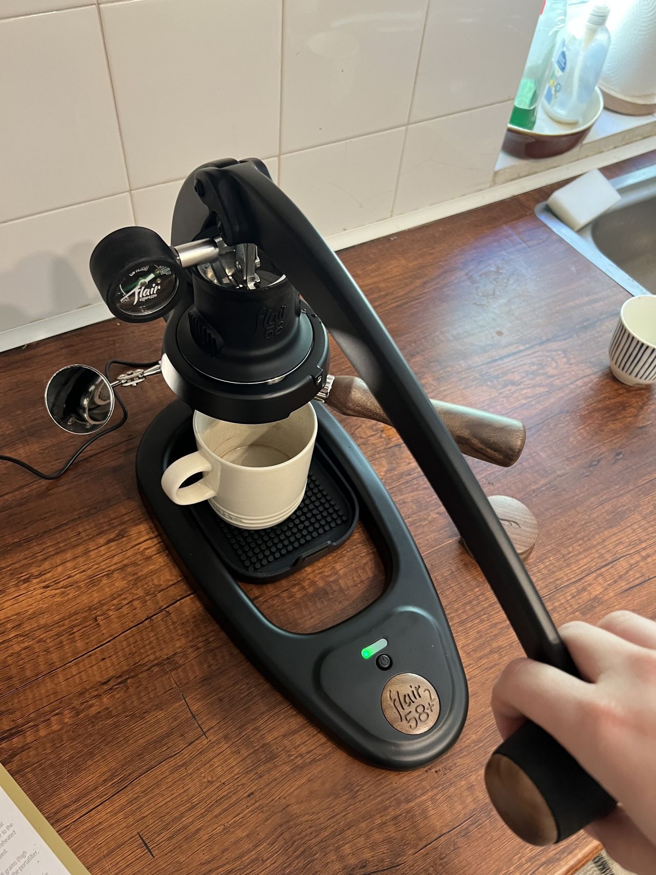 A white coffee mug under the brew head of a black and wood manual coffee machine sitting on a wooden countertop in a kitchen with white tiles on the wall.