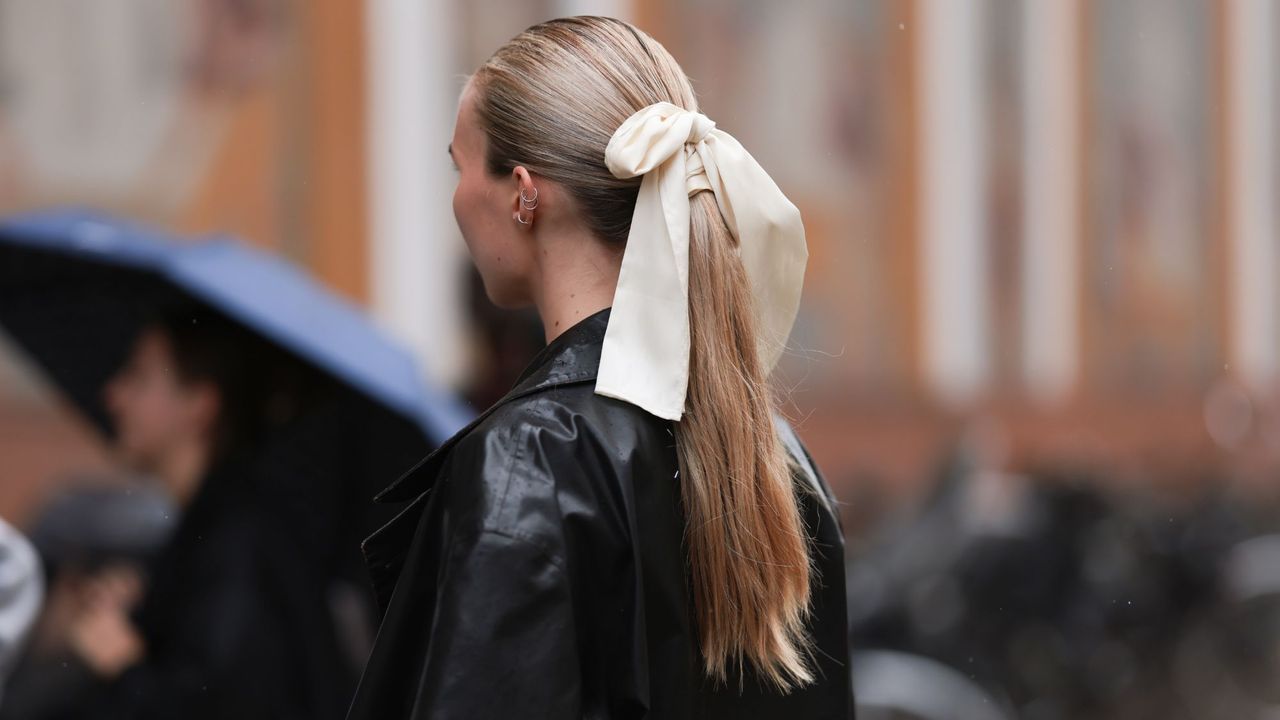 A guest at Copenhagen Fashion Week wearing silver earrings, cream white satin and silk ribbon as hair accessory, black leather long coat with a slicked ponytail and the best hairspray