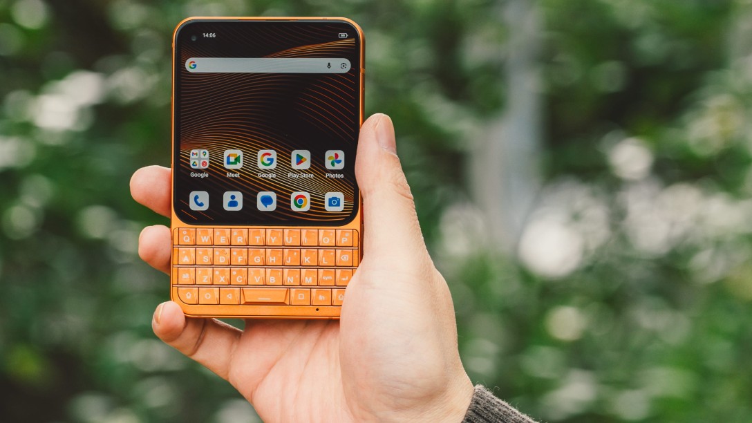 A person holds the orange Unihertz Titan 2 Elite upright against a blurred green leafy background, highlighting its compact size and physical keyboard.